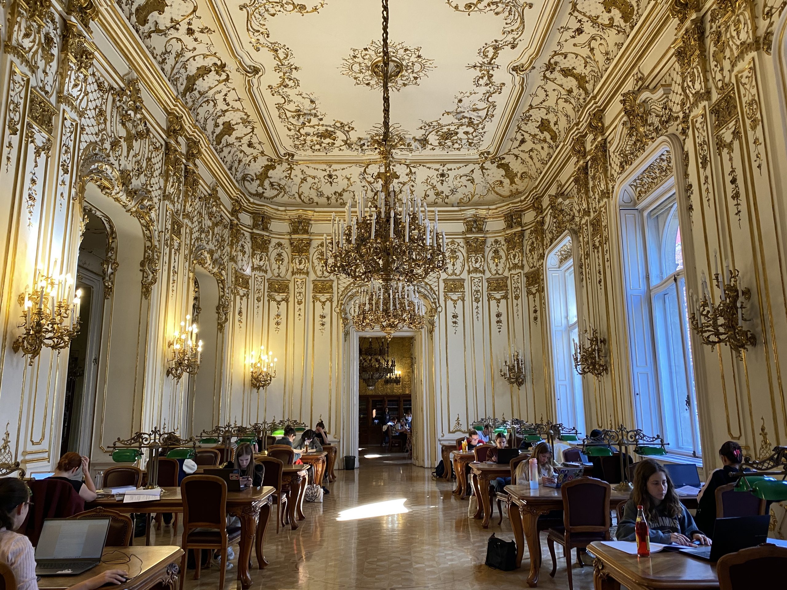 A ballroom with golden gilded walls and a golden chandelier filled with students studying at wooden tables